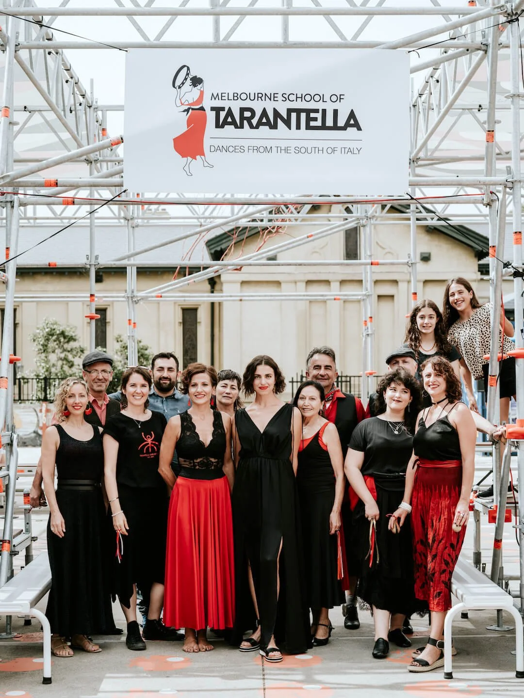 Group of people standing on stage with festival scaffolding and a sign that reads "Melbourne School of Tarantella - Dances from the South of Italy."