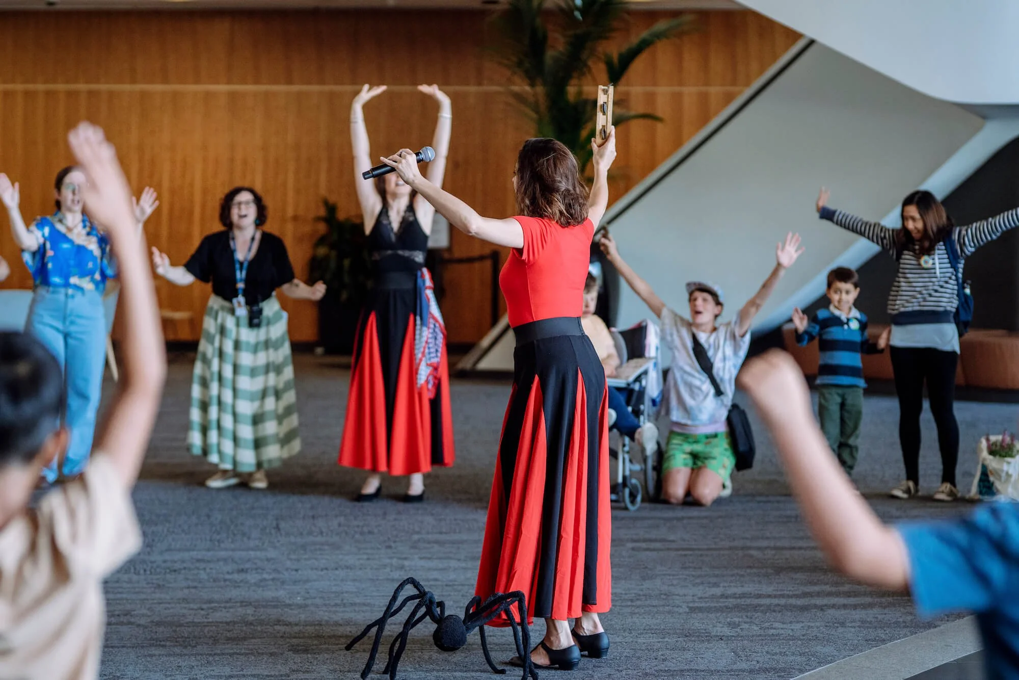 Women and children participating in a dance activity indoors, with a woman holding a microphone and a phone, wearing a red and black dress, with other women and children raising their hands in the background.