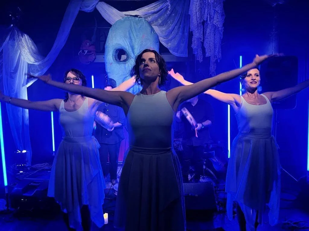 Three women on stage with arms outstretched, behind a large blue mask and draped fabric, illuminated by blue stage lights.