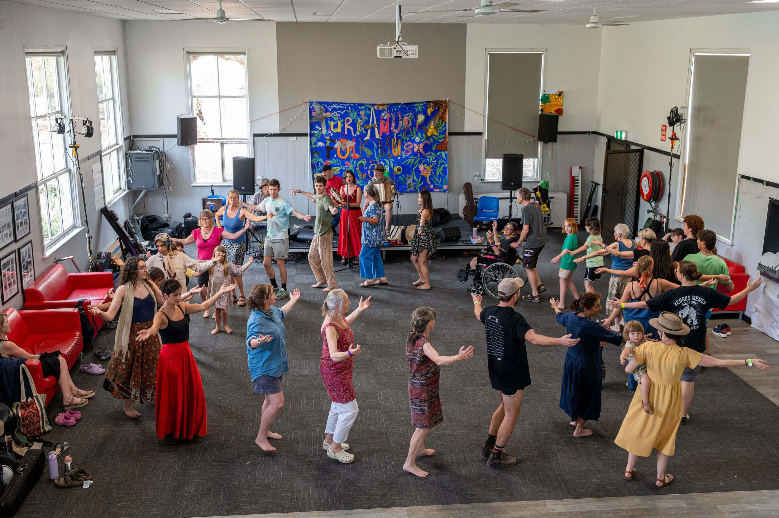 People of various ages dancing and participating in an indoor event with a colorful banner reading 'Turon, Amur, Follm, Music'. Some are standing, some are seated, and there is a person in a wheelchair. The room has large windows, sound equipment, and people seem engaged in a cultural or community celebration.