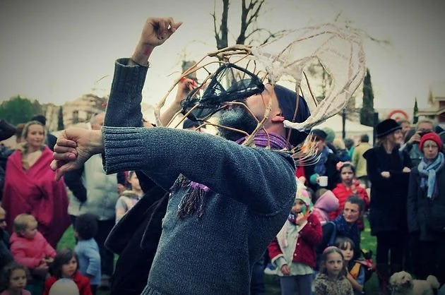 Person wearing a crown made of intertwined branches dancing at an outdoor event with children and adults in the background.