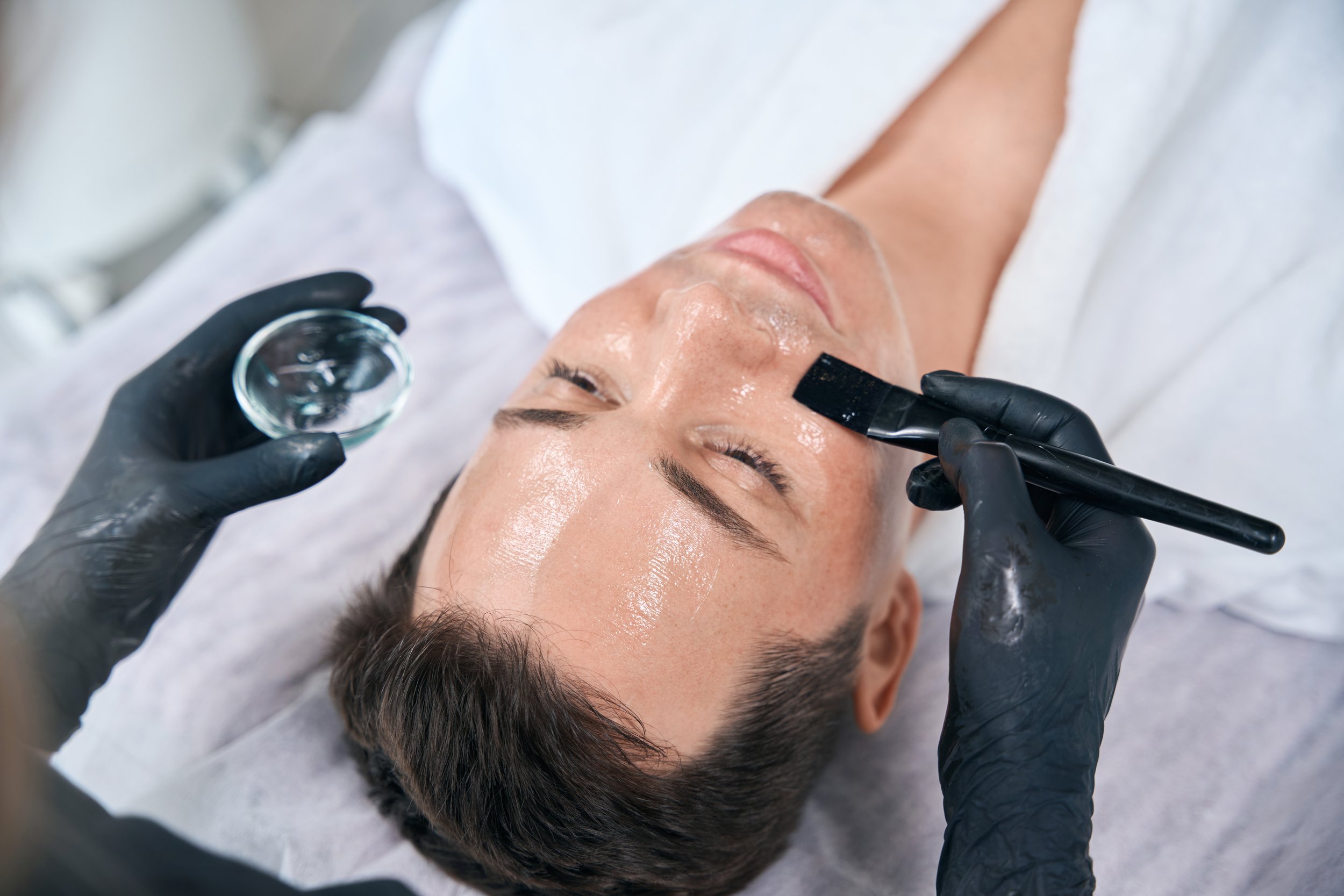 A woman lying on a treatment bed, getting a facial or skincare procedure with a laser device operated by a professional in a clinical setting.
