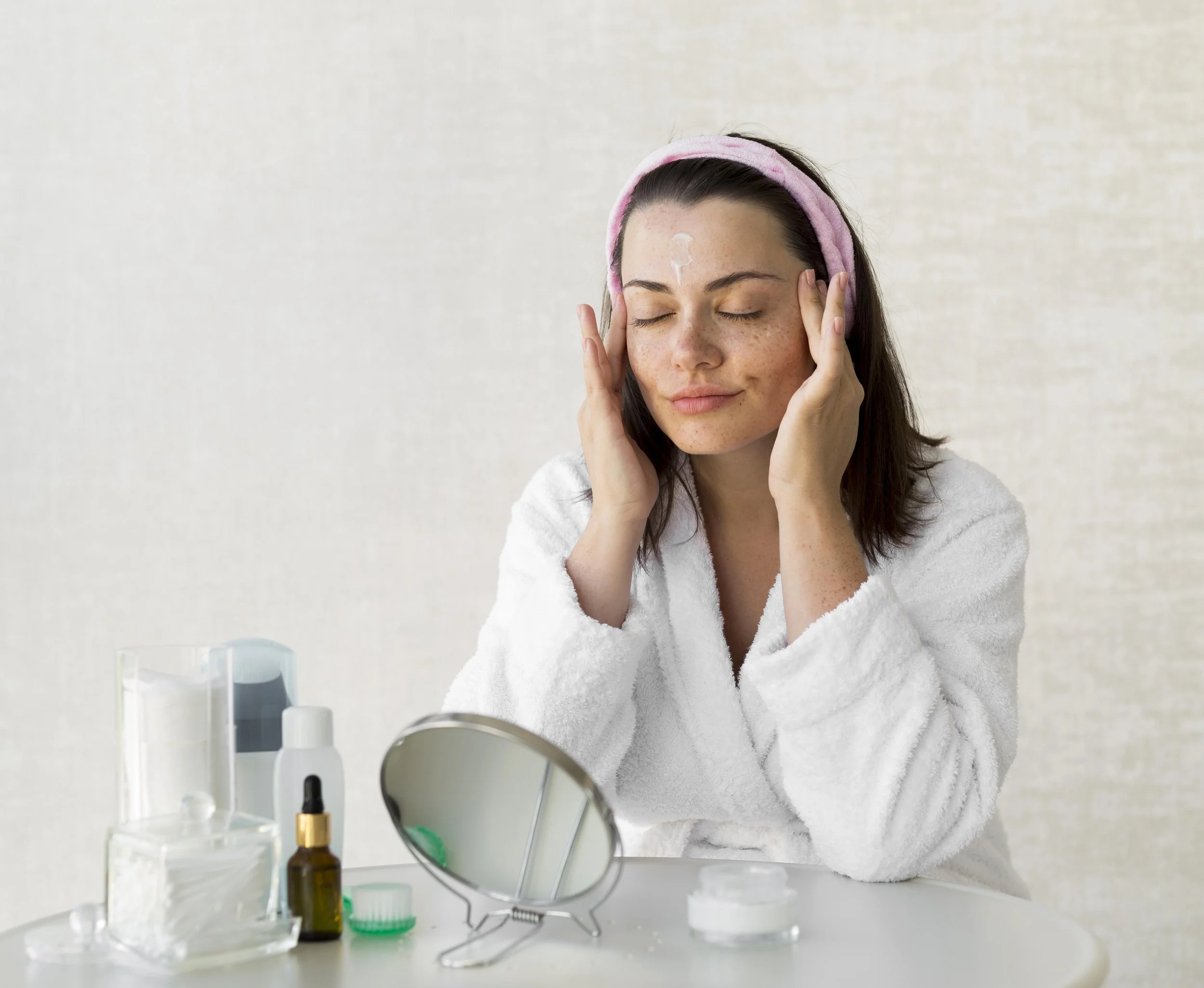 A woman applying skincare cream on her face in front of a mirror, surrounded by skincare products on a table.