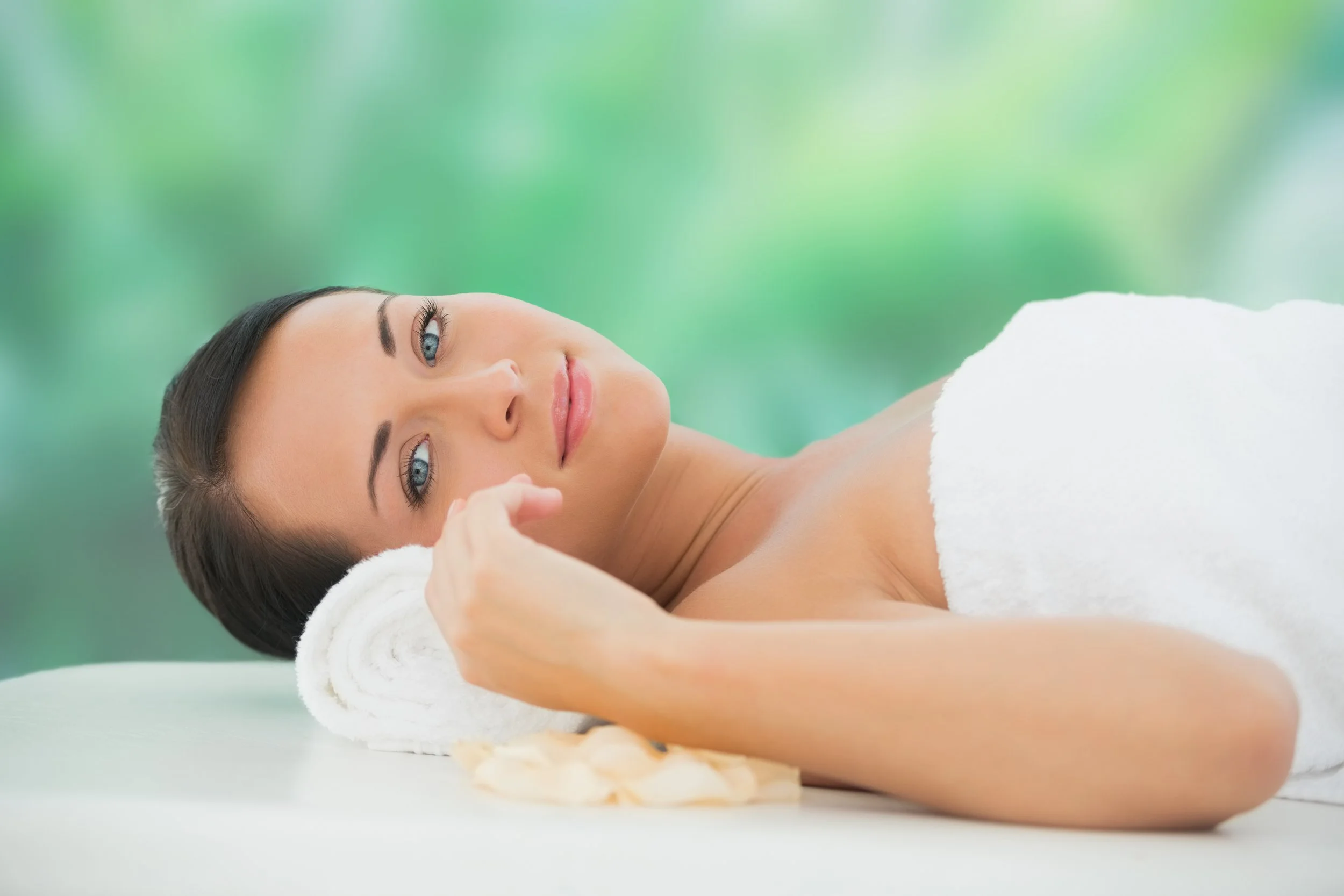 A woman lying on a massage bed with a white towel over her body, resting her head on a rolled towel, with a blurred green background.
