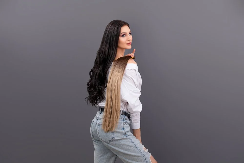 Young woman with long dark hair posing against a gray background, wearing a white off-shoulder blouse and high-waisted jeans.