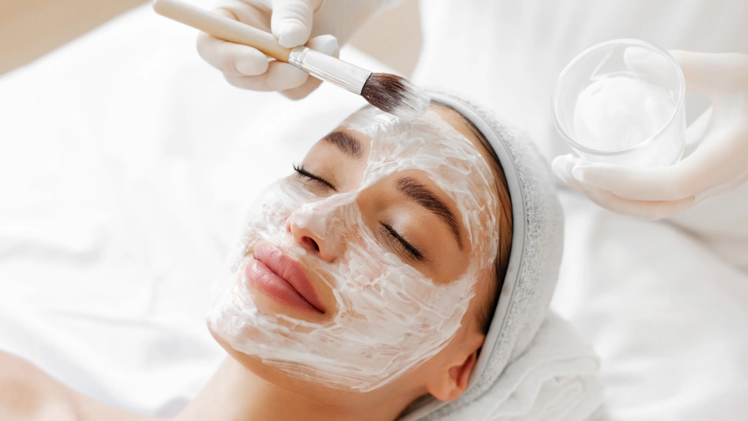A woman receiving a facial treatment, lying down with her eyes closed, as a skincare professional applies a cream or mask to her face with a brush, while holding a small glass of water.