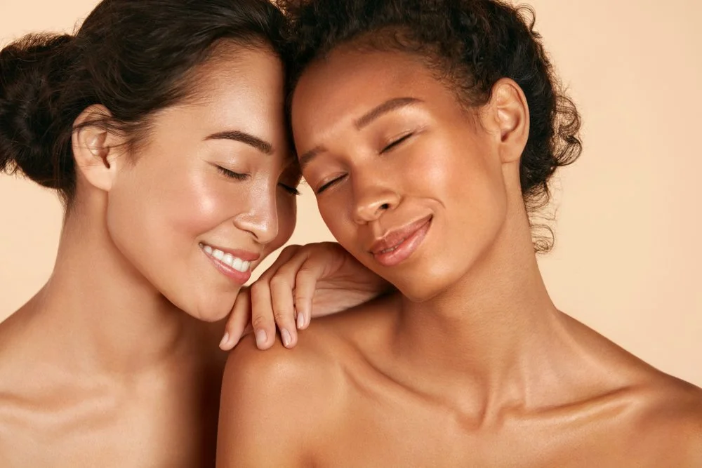 Two women with closed eyes smiling gently, touching shoulders, against a beige background.