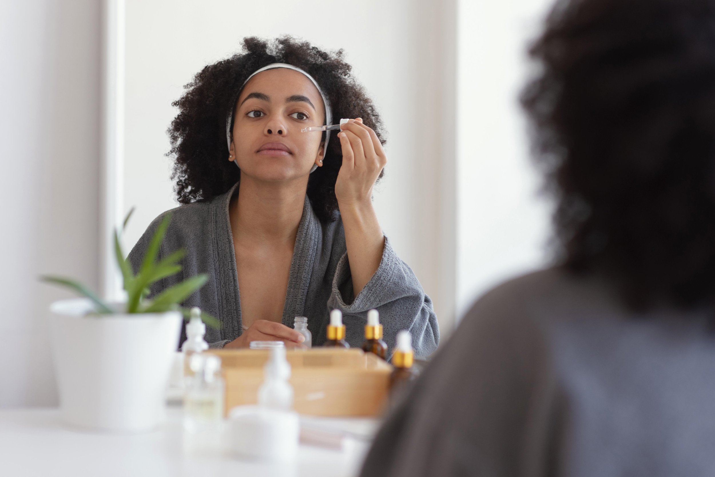 A woman with curly hair, wearing a headband and gray robe, applying skincare with a dropper while looking into a mirror.