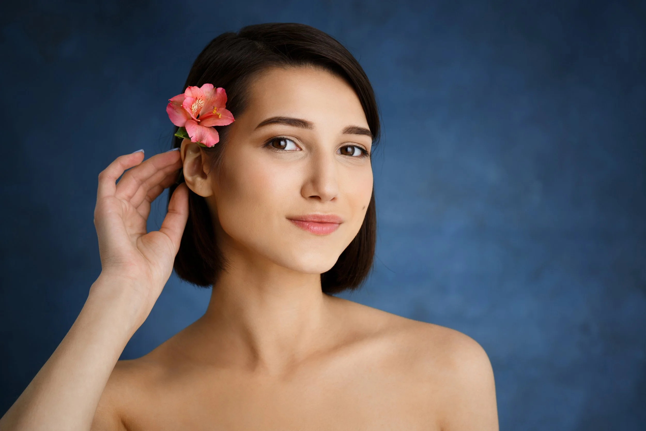 A woman with short brown hair and a pink flower in her hair against a dark blue background.