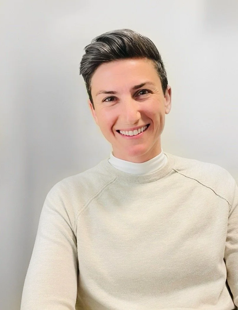 A smiling woman with short, dark, styled hair, wearing a beige sweater over a white turtleneck, standing against a plain light-colored background.