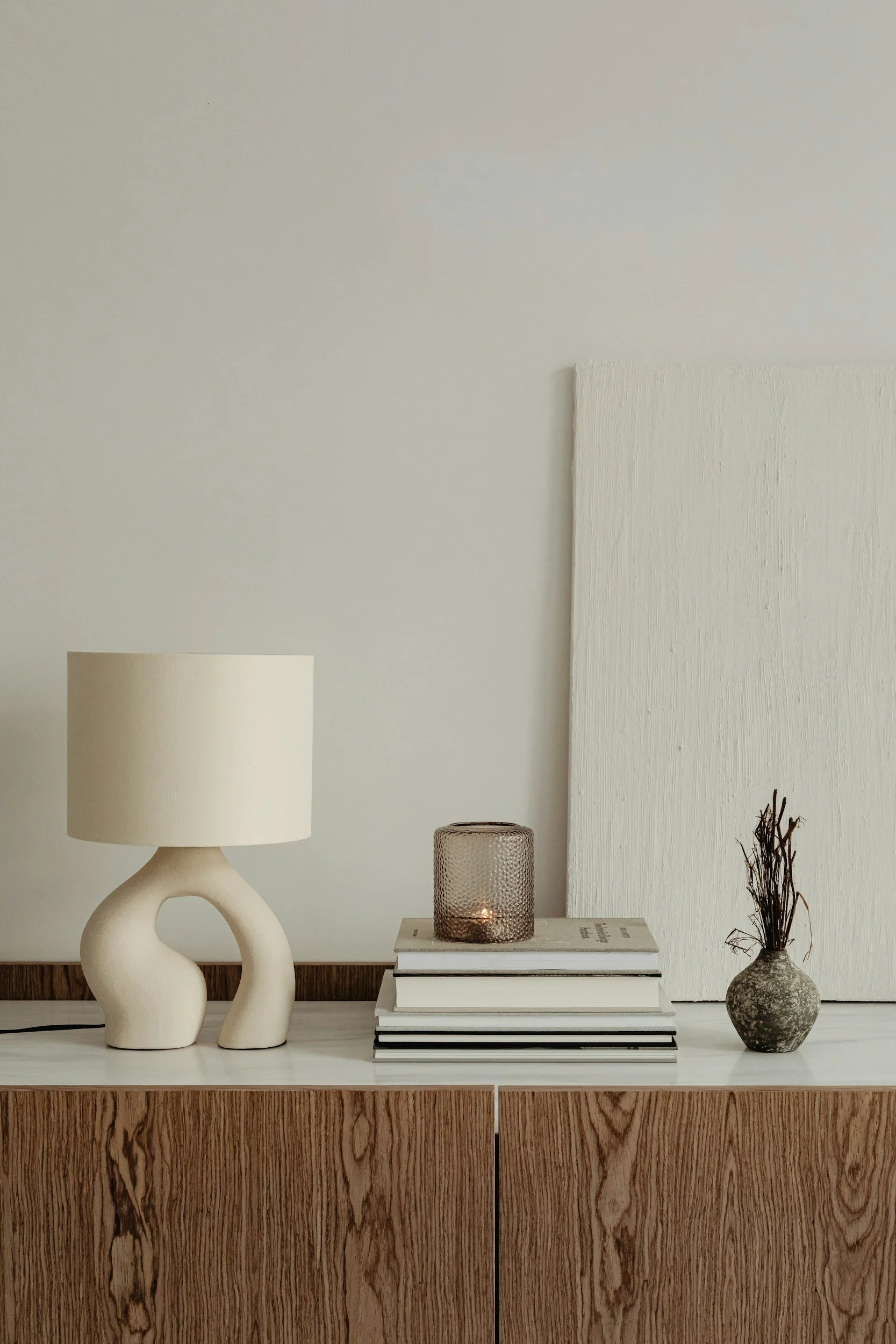 A minimalist interior decor with a white table top, a modern beige table lamp, a stack of books with a candleholder on top, and a small vase with dried twigs against a plain wall.