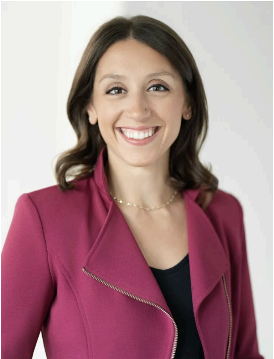 A woman with brown hair, smiling, wearing a pink blazer and a black top, standing against a plain light background.