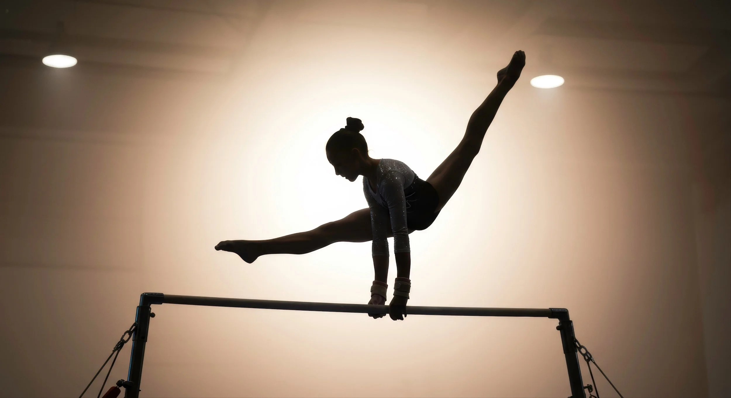 Silhouette of a gymnast performing an acrobatic move on a balance beam against a backlit wall.