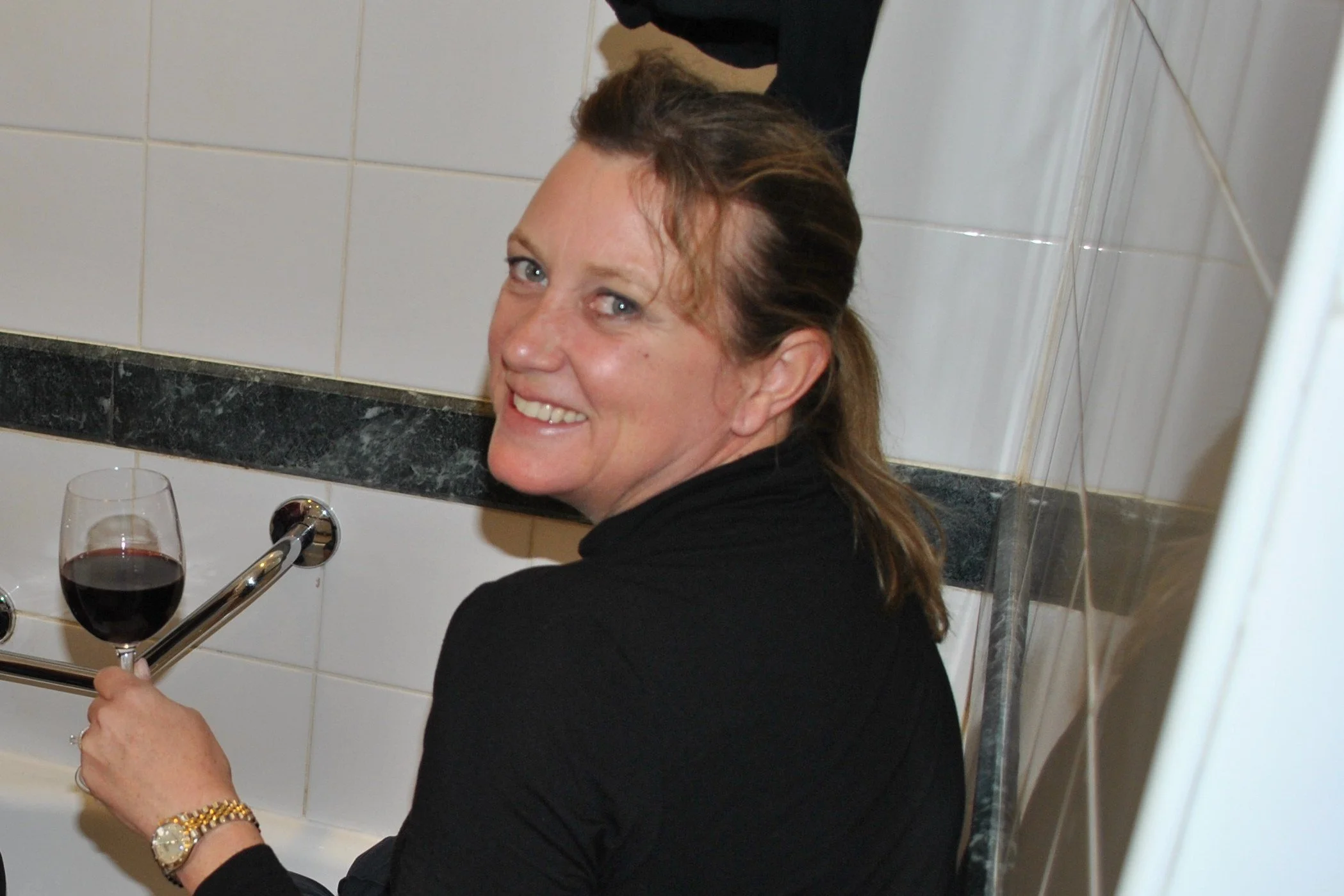 Brigitte perched on side of a bath, smiling over her shoulder with a glass of redwine
