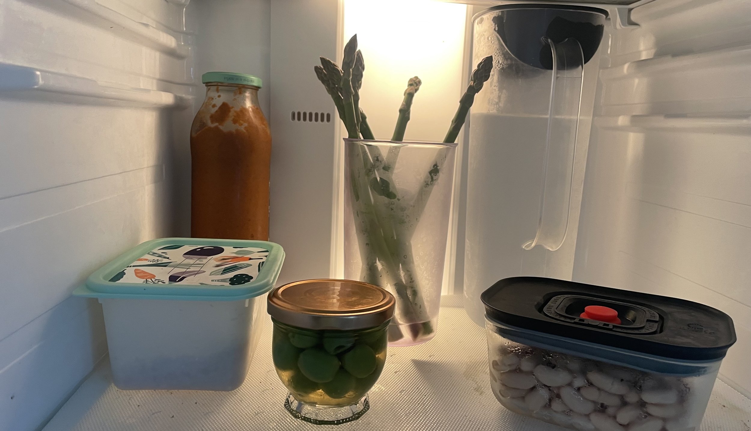 A small footed jar of green olives on a fridge shelf, backlit by the fridge light, beside asparagus standing in a glass, a jar of sauce and a container of white beans.