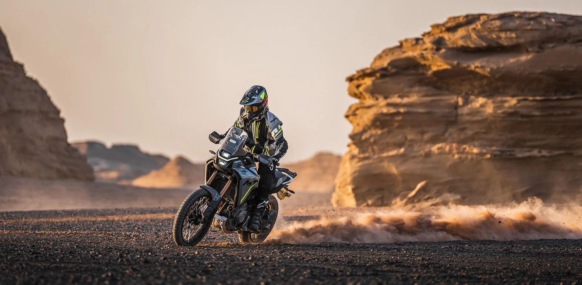 Motorcycle rider in black and yellow gear riding a dual-sport motorcycle across desert terrain with rock formations in the background during sunset.