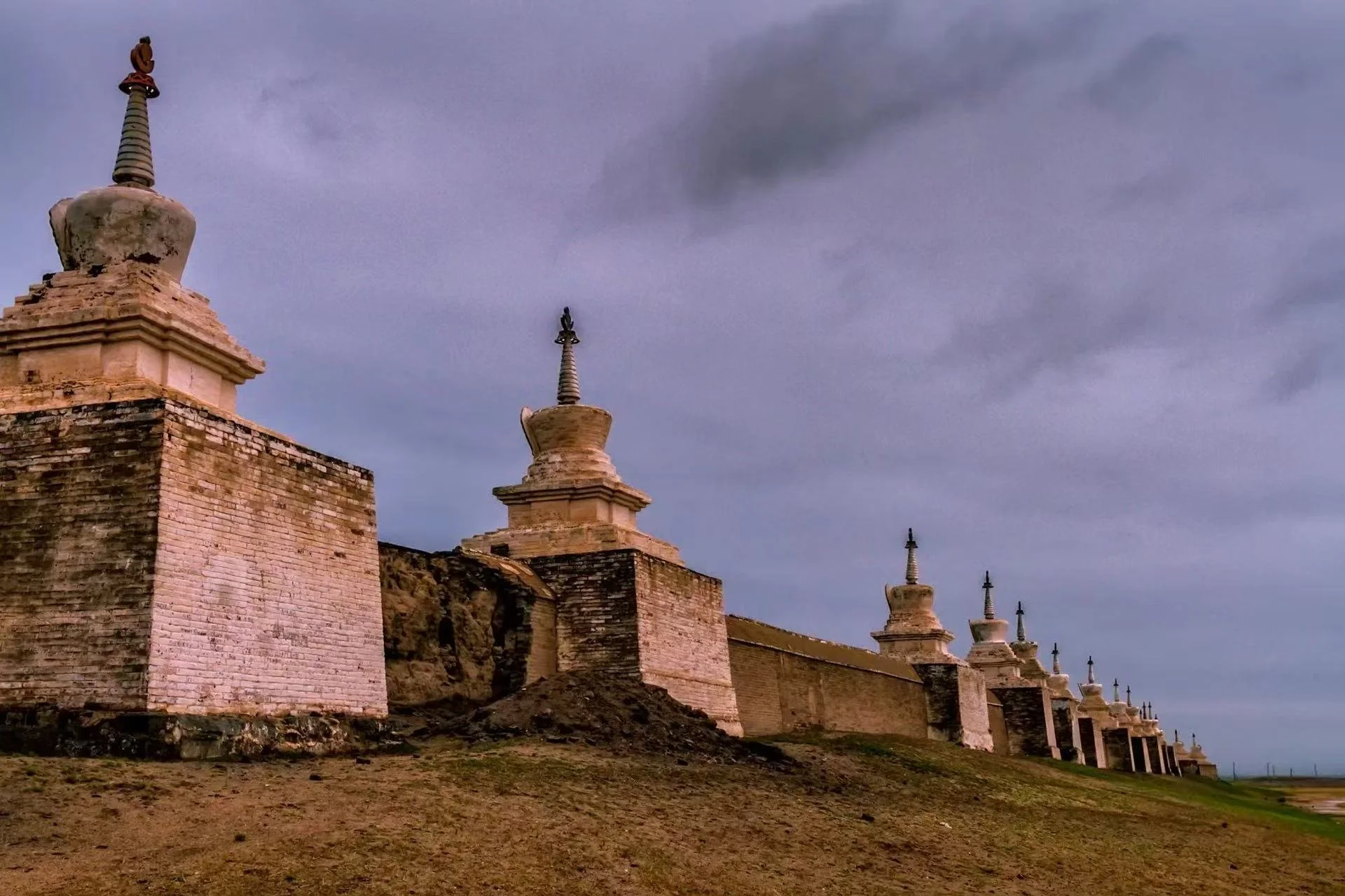 An ancient brick wall with several white stupas or pagodas on top, set against a cloudy sky. The wall runs along a grassy, rocky terrain.