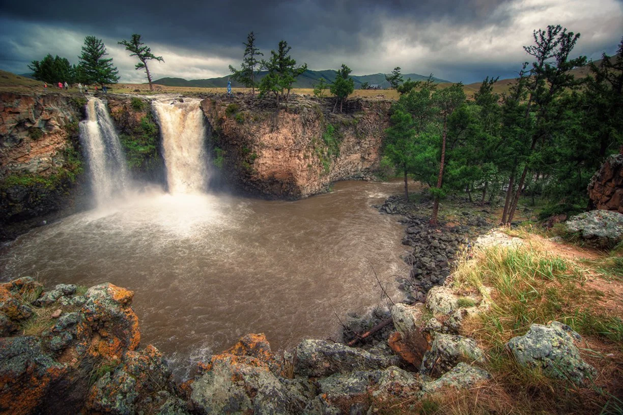 Two waterfalls cascading into a river in a rocky and forested landscape under a cloudy sky.
