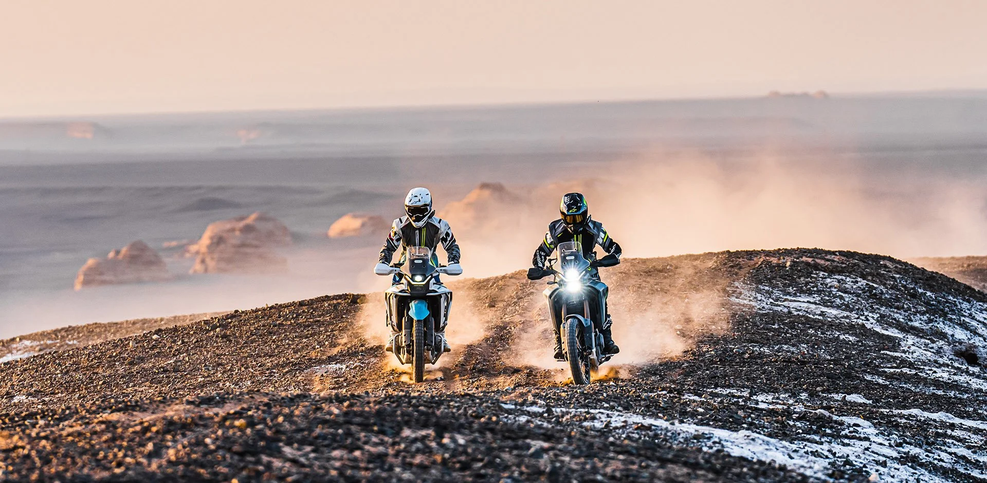 Two motorcyclists riding on a rocky desert terrain during sunset, dust and smoke rising behind them.