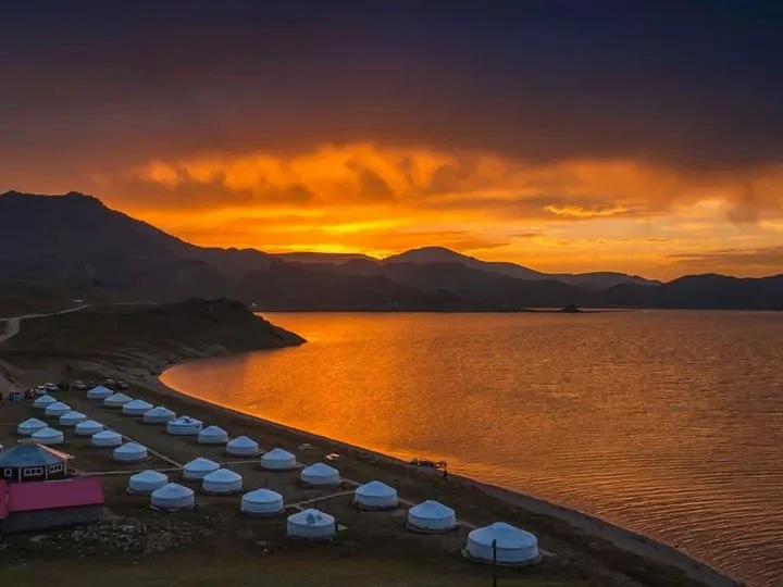 A lakeside scene at sunset with a vibrant orange sky and calm water reflecting the colors. In the foreground, there are numerous white yurts or tents arranged along the shore, and a pink building nearby. Hills and mountains are visible in the background.
