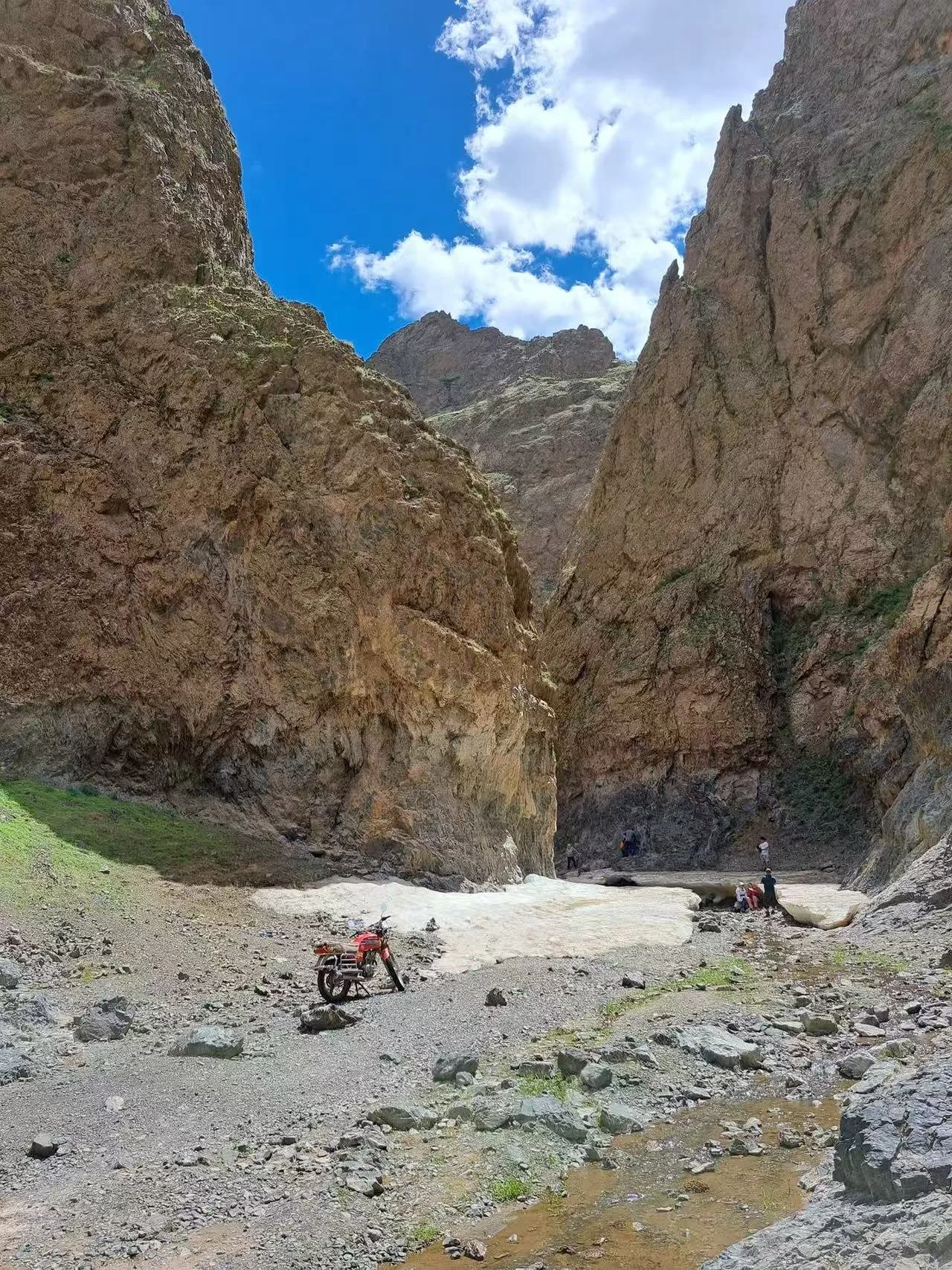 A motorcycle parked on rocky ground in a narrow canyon with tall, brown cliffs on either side and a bright blue sky with white clouds overhead.