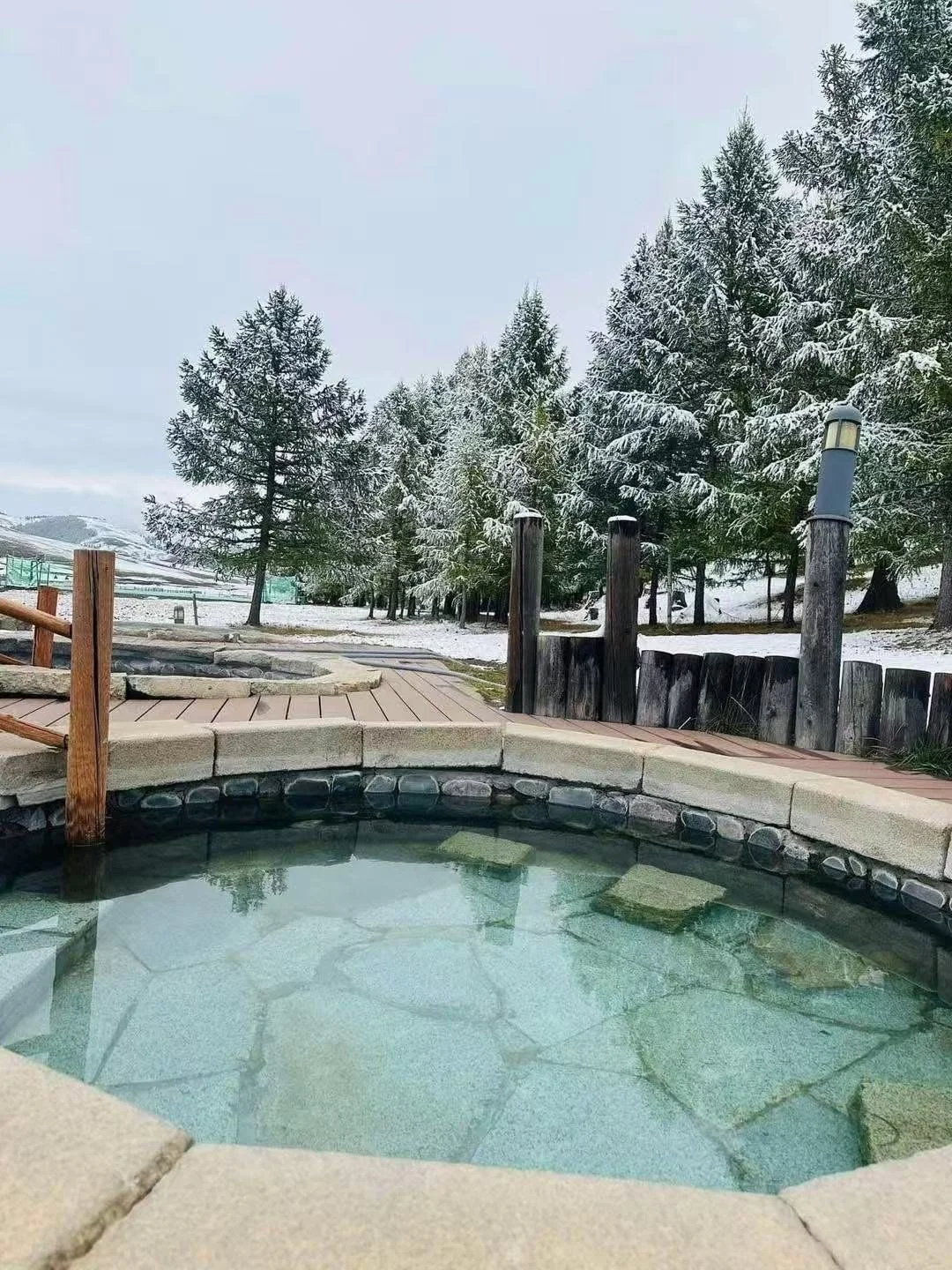 A hot spring pool or hot tub with stone and concrete border, set outdoors in a snowy landscape with snow-covered trees and a wooden pathway.
