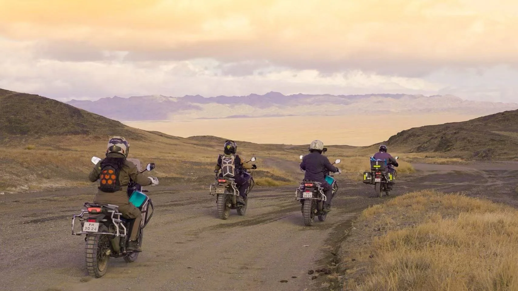 Four motorcyclists riding off-road on a dirt path in a barren, hilly landscape with mountains in the distance under a cloudy sky.