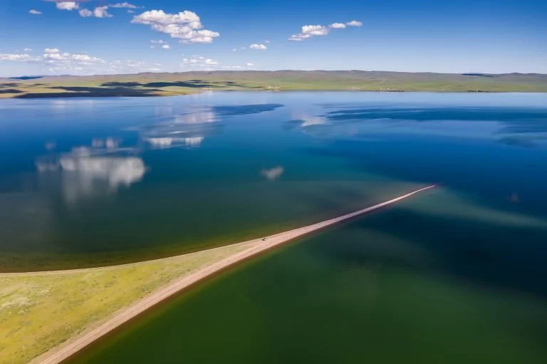 A large body of water with a long, narrow dirt road extending into it, under a partly cloudy sky with distant green hills.