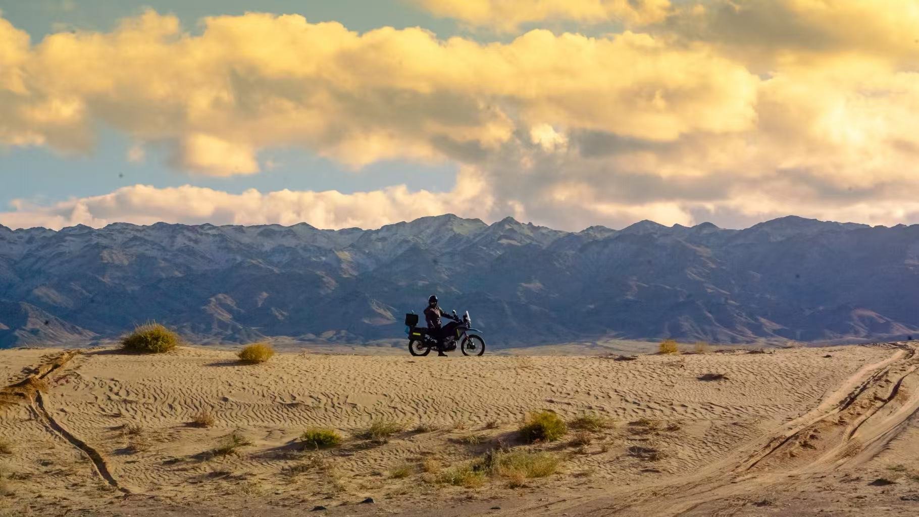 A person riding a motorcycle in a desert landscape with mountains in the background and clouds in the sky.