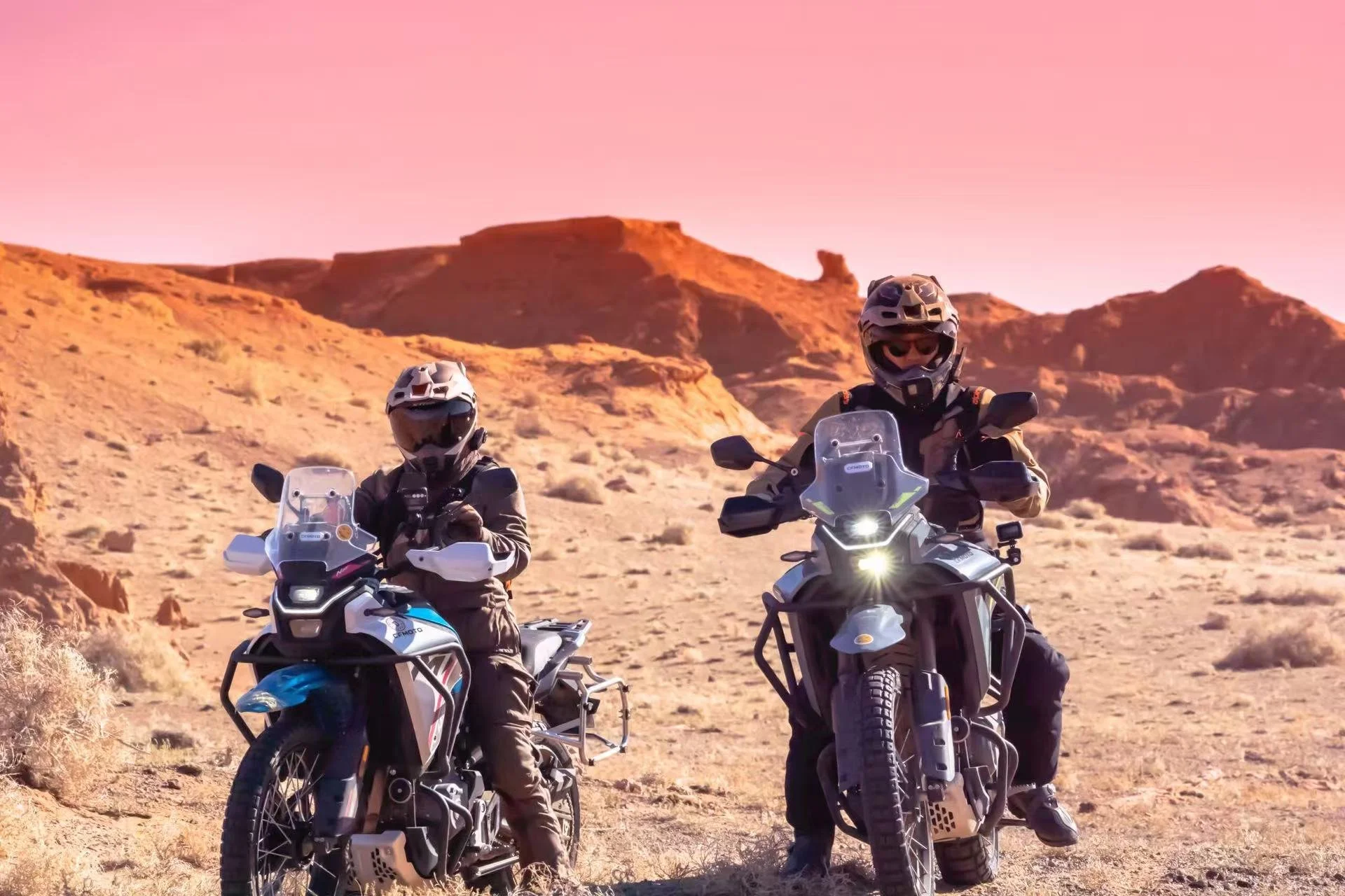Two motorcyclists in riding gear and helmets riding adventure motorcycles through a desert landscape with rocky hills and a pink sky.