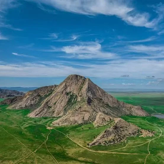 Distant view of a large rocky mountain with green plains and trails at the base, under a mostly clear blue sky with scattered clouds.