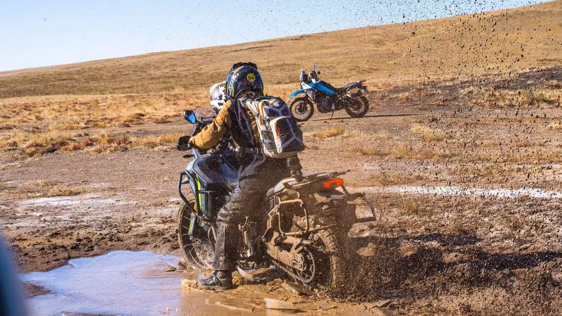 Motorcycle rider crossing a muddy puddle in a desert landscape with another motorcycle in the background.