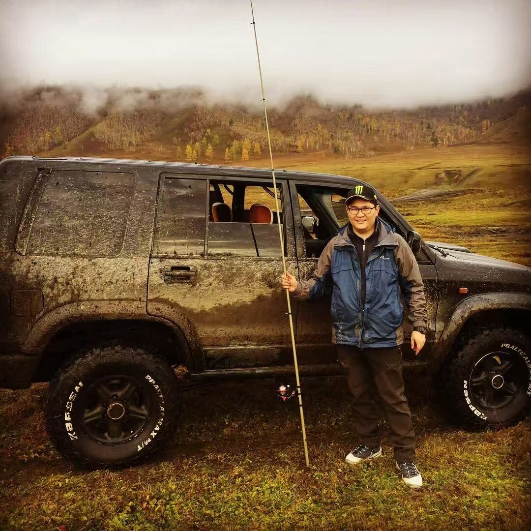 A man standing next to a muddy black SUV on a grassy terrain with mountains and cloudy sky in the background; the man is holding a long stick, wearing a blue and gray jacket, black pants, sneakers, glasses, and a Monster Energy cap.