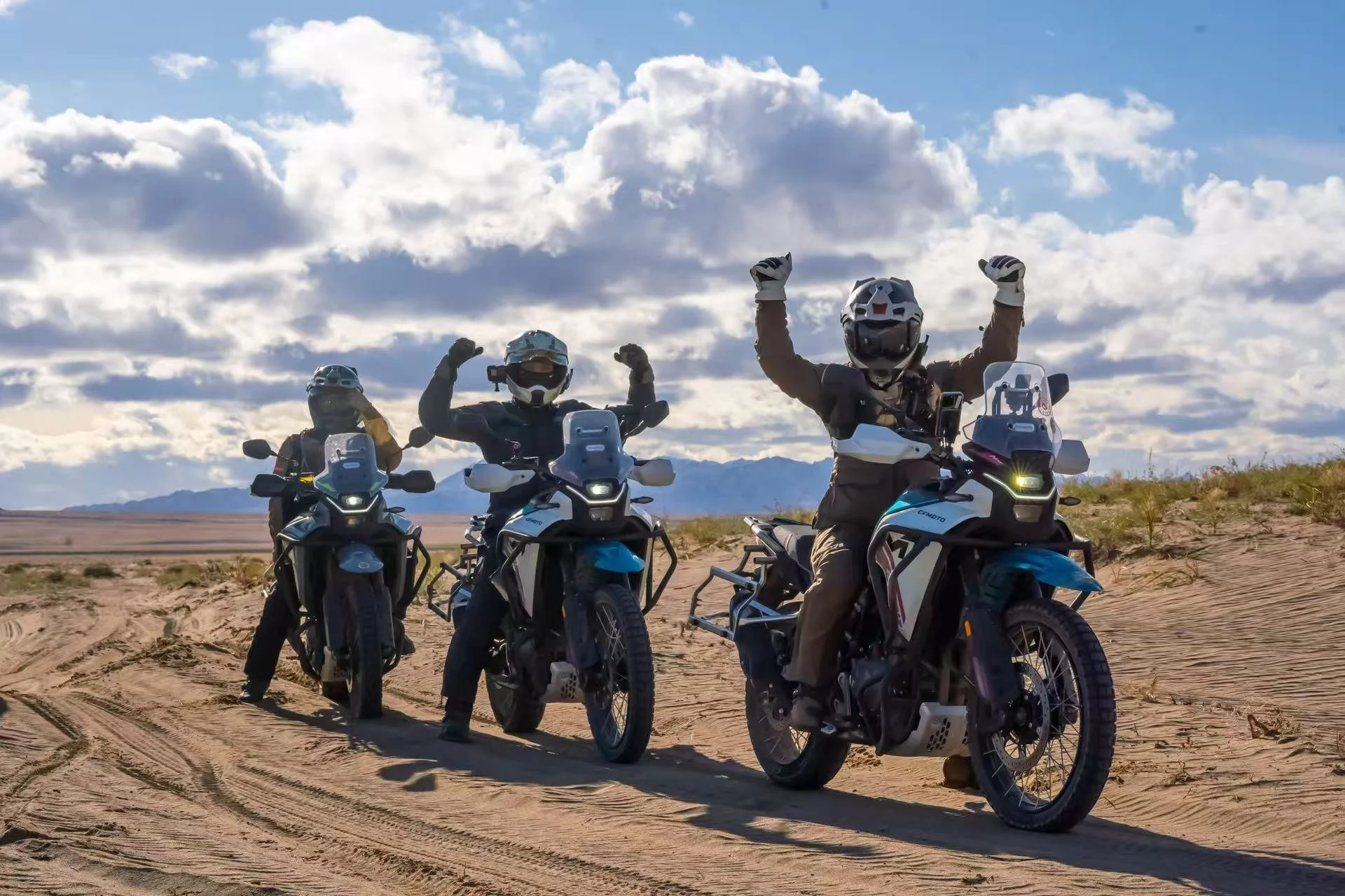 Three motorcyclists riding adventure motorcycles through a desert landscape with mountains in the distance, under a partly cloudy sky.