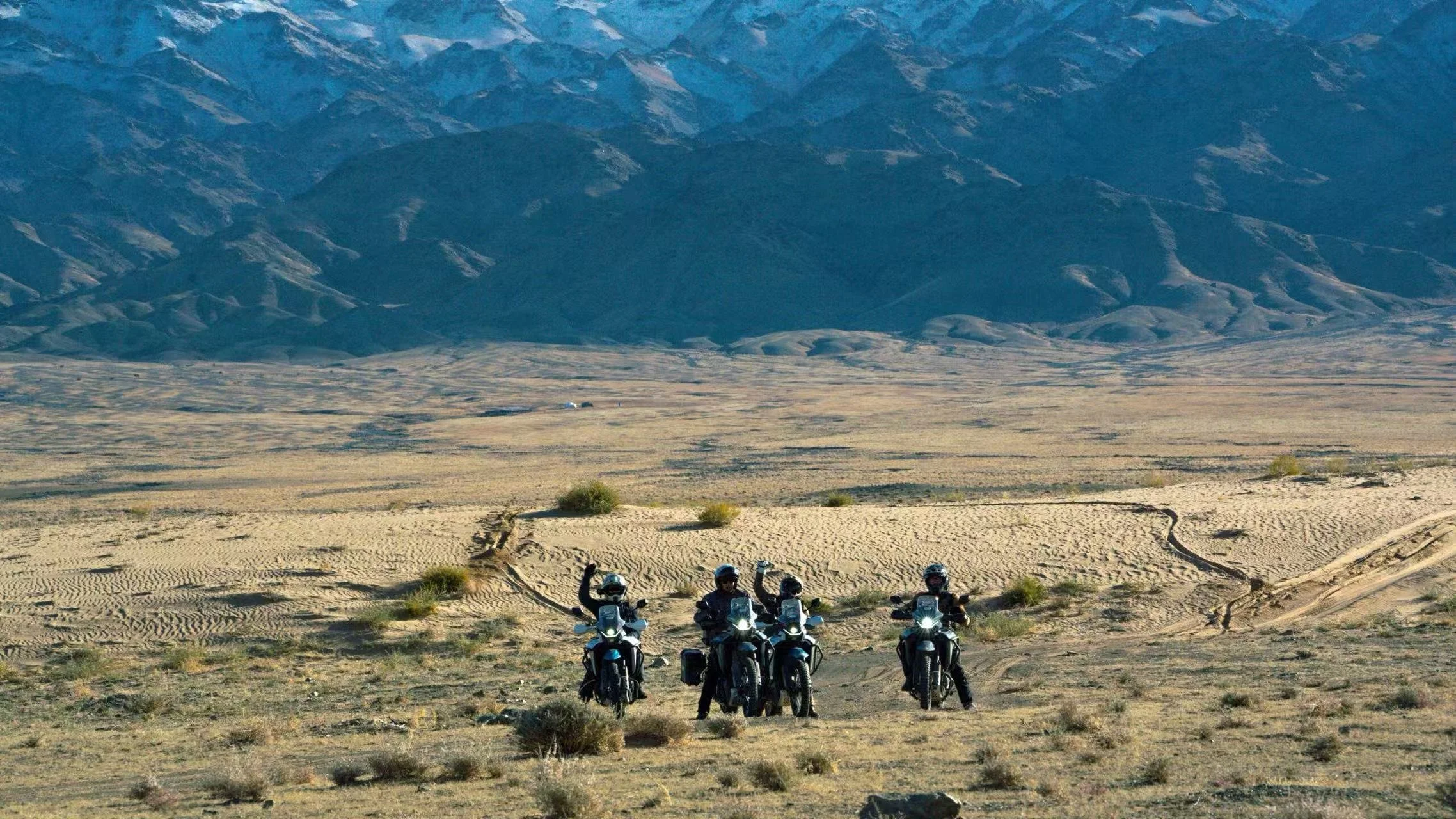 Four motorcyclists in gear, riding through a desert landscape with mountains in the background, some riders waving.