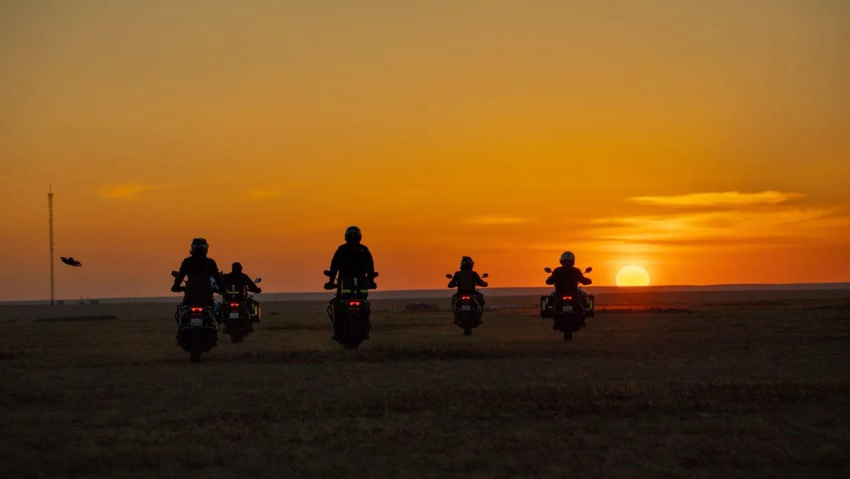 Five people riding motorcycles at sunset on a flat landscape with a tall communication tower on the left.