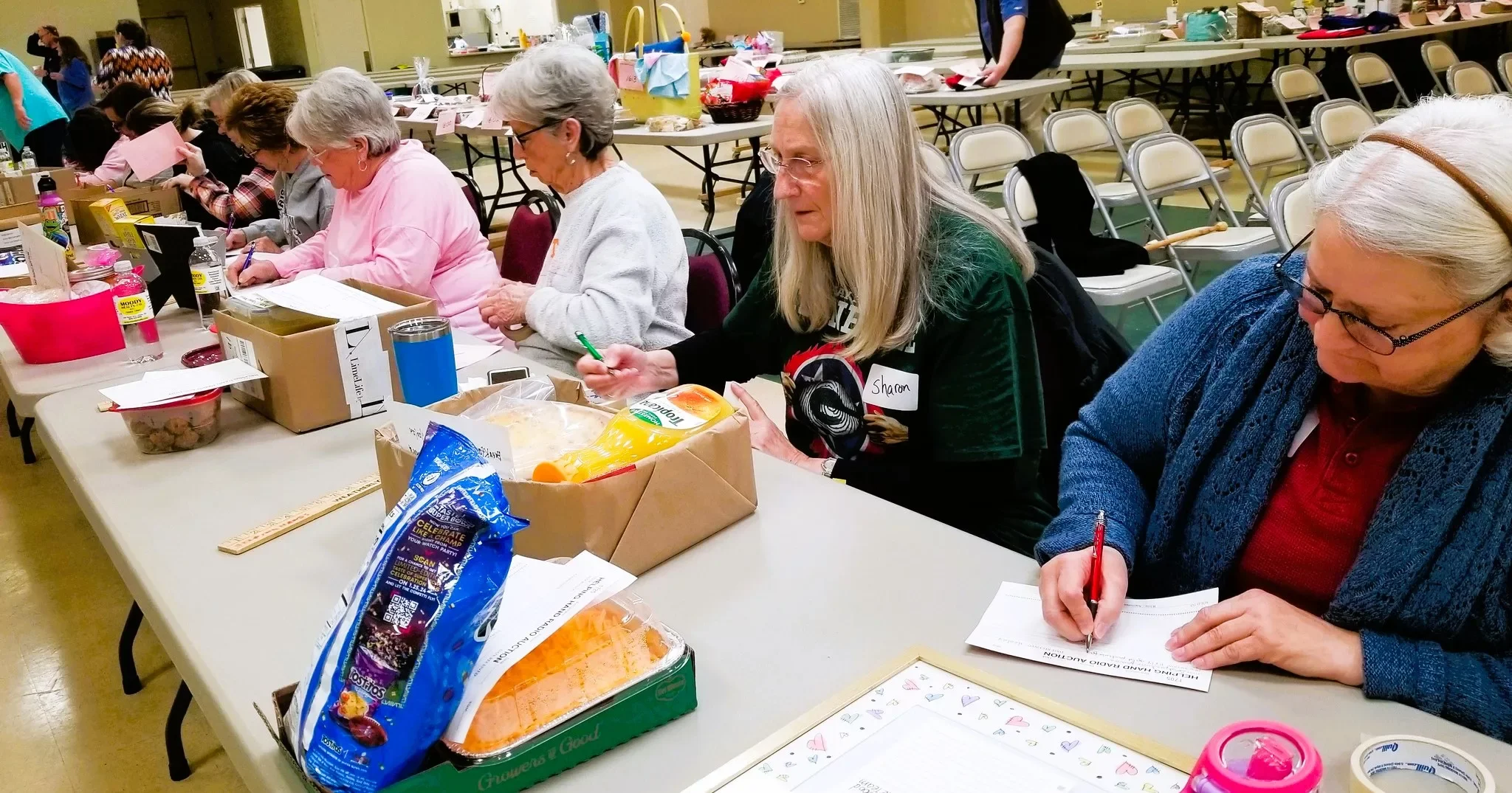 A group of elderly women sitting at a long table, writing and taking notes, with food and supplies on the table in a community or event setting.