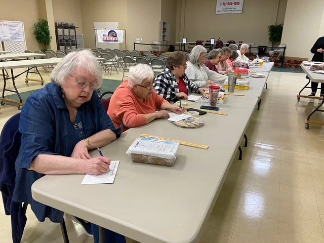 Older women sitting at a long table in a community center, working on crafts or projects, with supplies and rulers in front of them.