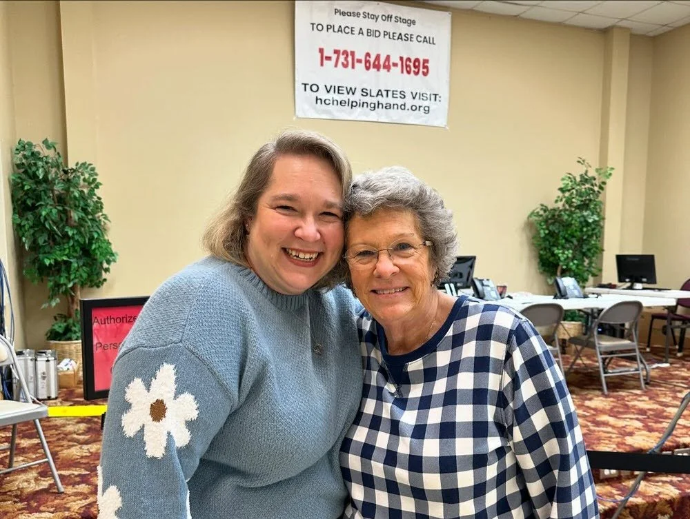 Two women smiling and standing close together in an indoor setting, with a sign and several chairs and tables in the background.