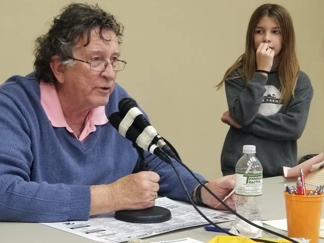An elderly man with glasses and a woman with long brown hair listening at a table during a discussion or meeting.