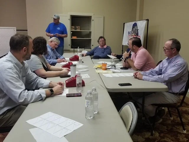 A group of people sitting around a conference table in a meeting room, with one person standing near a cabinet and a presentation board.