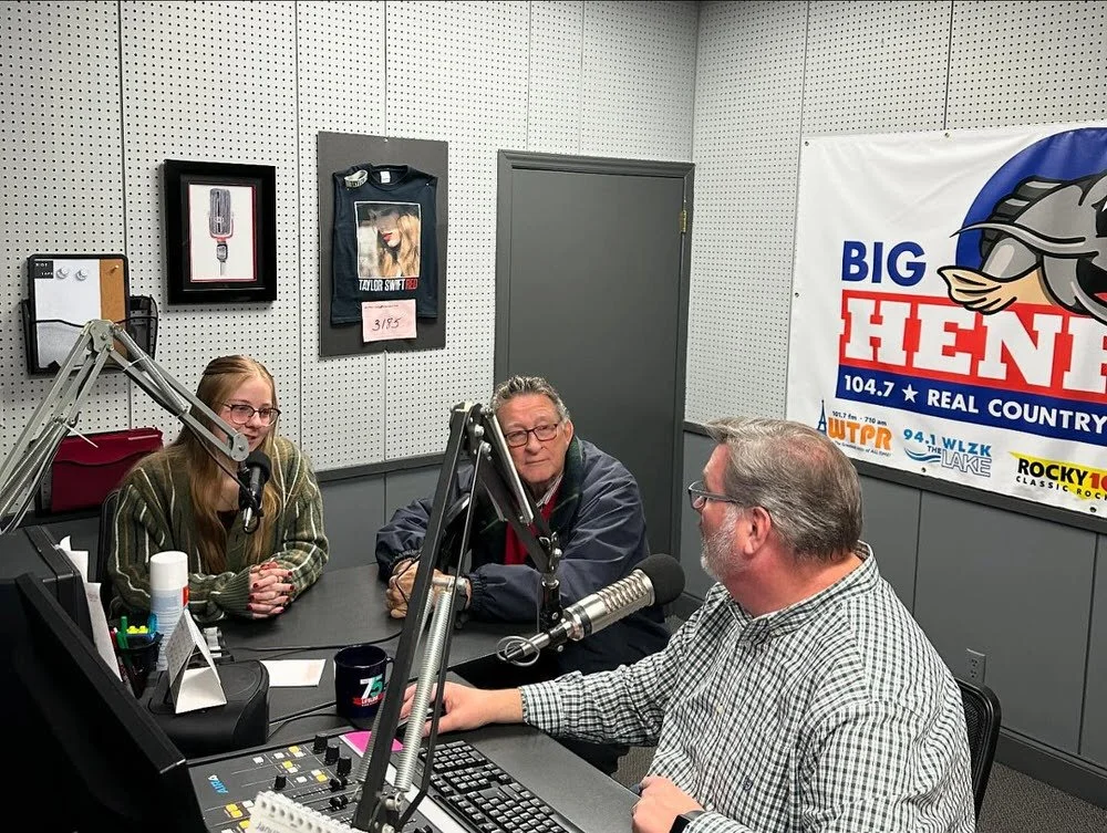Three people sitting at a radio station table with microphones, in a studio with Big Hank logo on the wall.