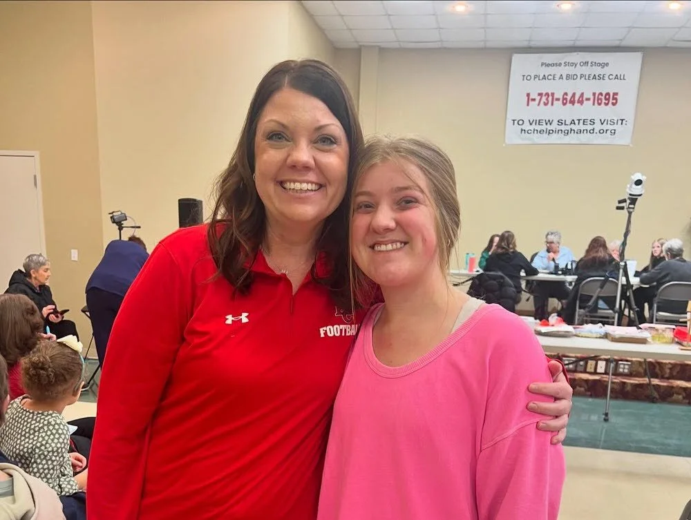 Two smiling women, one in a red sports shirt and the other in a pink sweatshirt, standing close together in an indoor setting with a group of people seated at a table in the background.