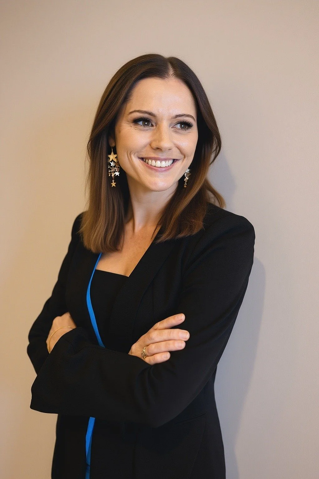 A woman with shoulder-length brown hair smiling and wearing star-shaped earrings, dressed in a black blazer and standing against a beige wall.