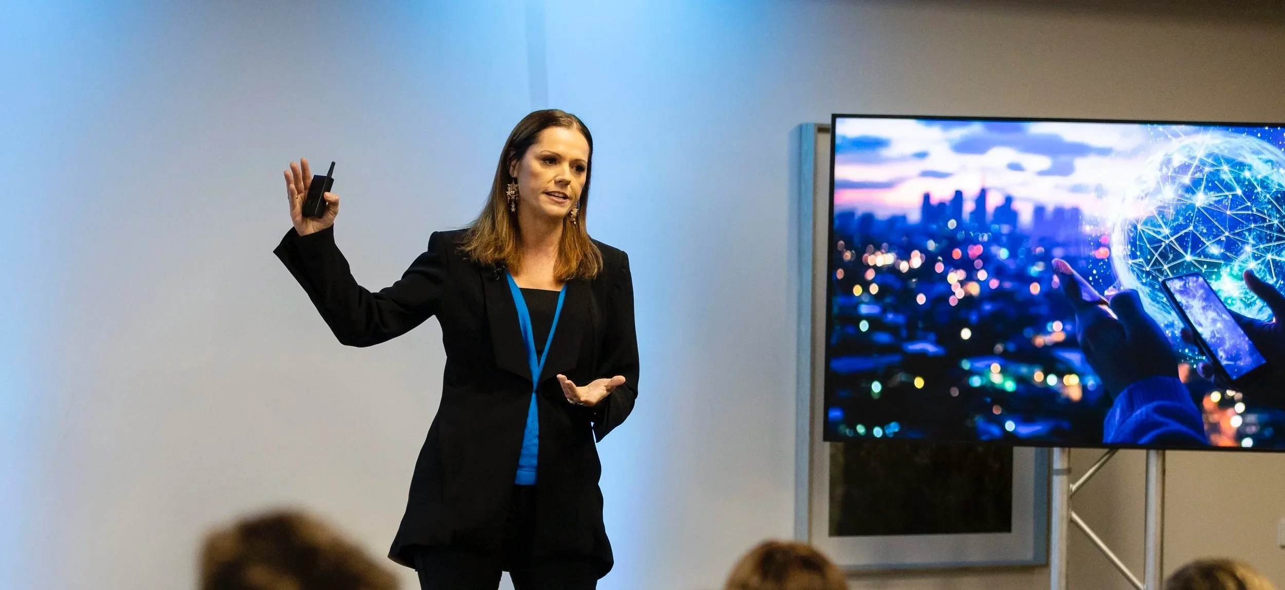 Rebecca Maklad - The Human Edge | Speaker A woman in a black blazer and earrings is giving a presentation, holding a clicker in her right hand and making a gesture with her left hand.