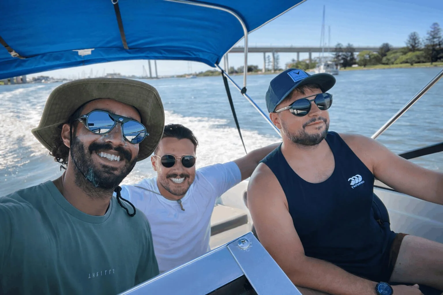 Three men smiling and enjoying a boat ride on a sunny day with water and bridge in the background, all wearing sunglasses.