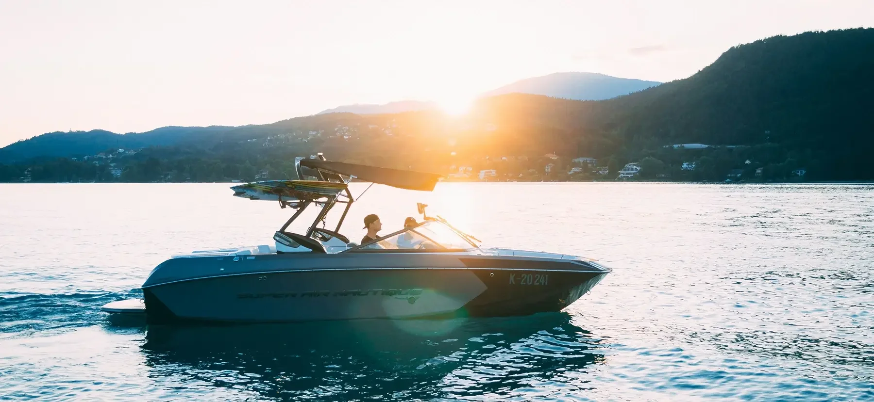 A motorboat with two people aboard, cruising on a calm lake during sunset, with mountains and houses in the background.