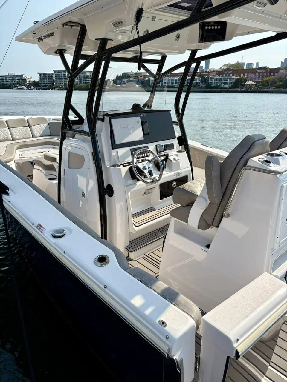 View of the cockpit and seating area of a luxury motorboat on a calm waterway with modern residential buildings in the background.