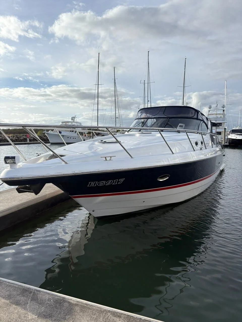 A white and blue yacht docked at a marina with other boats in the background, under a cloudy sky.