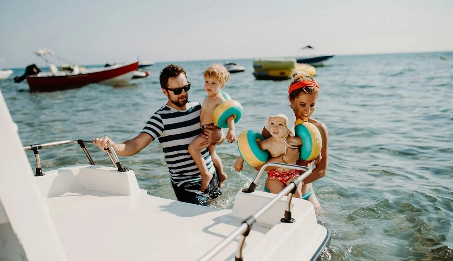Family of four in swimwear at the beach, children with flotation devices, arriving by boat, with boats and the sea in the background.